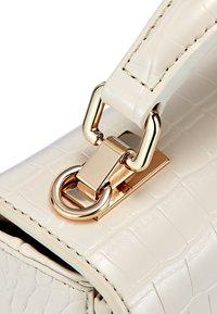 Kremowa torba o fakturze krokodylej, z metalowym zapięciem w złotym kolorze i akcesoriami. Gładki skórzany pasek z widocznym przeszyciem.