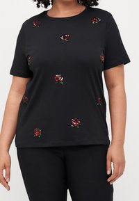 Svart kortärmad topp med spridda röda paljettrosdekorationer, bärs av en person med lockigt hår, tillsammans med svarta byxor.