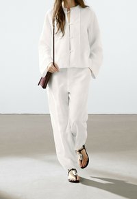 White linen top with a cropped design, featuring a keyhole opening and button accents, paired with matching loose fitting trousers and black sandals.