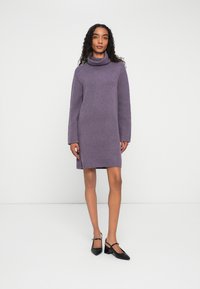Frau mit langen lockigen Haaren, die ein lila Rollkragenpulloverkleid und schwarze, niedrige, spitz zulaufende Schuhe trägt, und vor einem schlichten weißen Hintergrund steht.