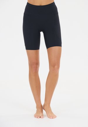 Kvinde iført sorte, højtaljede, tætsiddende biker-shorts, står barfodet på en hvid baggrund.