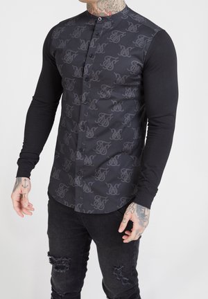 Mand iført sort slim-fit skjorte med mønstret frontpanel, ensfarvede lange ærmer og revnede sorte jeans, der viser tatoverede hænder og nakke.