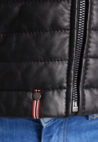 Černá kožená bunda s prošívanou texturou, stříbrný zip a malá visačka s červenými, bílými a černými pruhy.