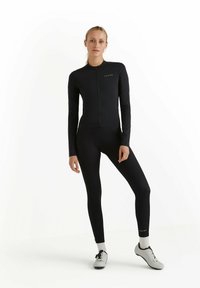 Pantalon de cyclisme noir à manches longues avec fermeture éclair intégrale, présentant un design épuré et une coupe ajustée. Accentué par un texte vert et associé à des chaussures grises.