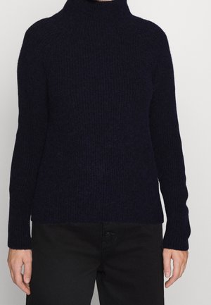 Persoon draagt een aangesloten zwarte geribbelde coltrui en zwarte broek, staand tegen een effen lichte achtergrond.