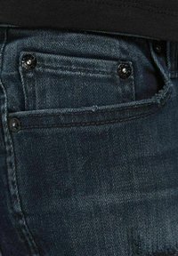 Detailní záběr na kapsu tmavě modrých džínů s kovovými nýty a část černé košile viditelná nad pasovým lemem.