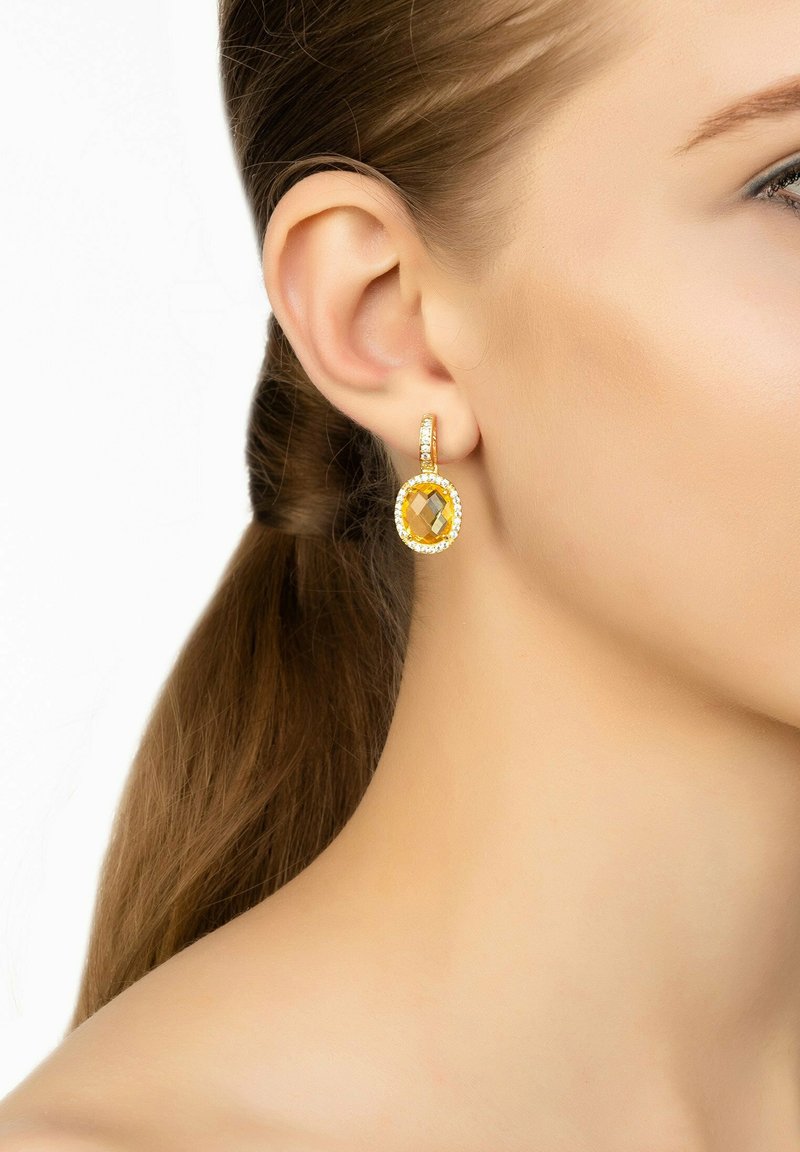 Guldörhängen med en stor gul kristall i mitten, omgiven av små klara stenar, fästa med en liten hooplås.