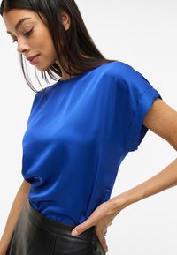 Top in satin blu con maniche corte, scollatura rotonda e texture liscia. Abbinato a una gonna in pelle nera, evidenziando il contrasto nei materiali.