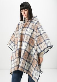Woman wearing a beige, white, and navy plaid hooded poncho over a light sweater and blue jeans, standing against a white background.
