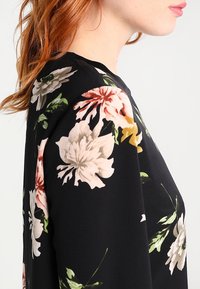 Schwarze Bluse mit floralem Muster, die große rosa, weiße und grüne Blumen zeigt. Glatter Stoff mit lockerer Passform, Rundhalsausschnitt und langen Ärmeln.
