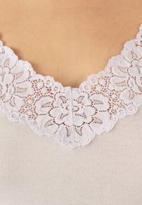 Top beige con un escote de encaje blanco ondulado, que presenta patrones florales y una textura acanalada en la tela.