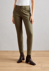 Olivově zelené slim-fit kalhoty s hladkou texturou, se dvěma předními knoflíky a bočními kapsami, doplněné černými kotníkovými botami na podpatku.