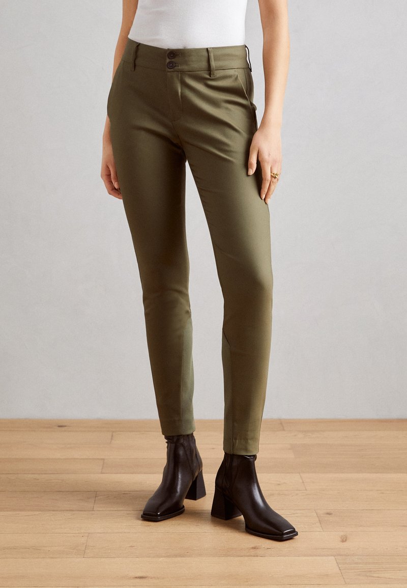 Olivově zelené slim-fit kalhoty s hladkou texturou, se dvěma předními knoflíky a bočními kapsami, doplněné černými kotníkovými botami na podpatku.