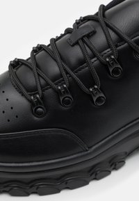 Svart skinnsko med strukturerad yta, som har elastiska snören, sex låsningsmekanismer och en tjock gummisula med cirkulära mönster.