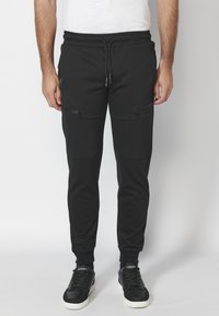 Černé jogger kalhoty vyrobené z měkké tkaniny, s elastickým pasem na šňůrku, obsahující kapsy na zip a úzké manžety na kotnících.