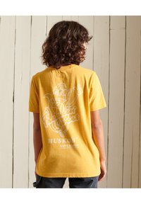 Camiseta de algodón amarilla con estampado gráfico blanco en la parte posterior que incluye texto y un diseño de dibujo animado. Mangas cortas y un corte relajado.