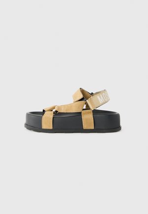 Sandalias de ante beige con suelas de goma negras, que cuentan con correas ajustables y una tira con marca en la parte trasera. Diseño plano con un acolchado mínimo.