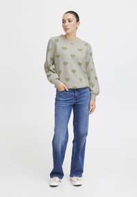 Pull gris clair avec des motifs de cœur verts, col rond et coupe décontractée, associé à un jean bleu et des baskets blanches.