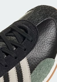 Black leather sneaker with textured green accents, featuring a mesh overlay, white stripes, and black lace-up detail. Rubber outsole.