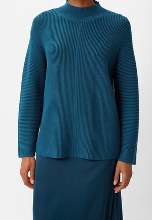 Pull en maille côtelée sarcelle avec col montant et manches longues porté avec une jupe assortie sarcelle, présenté des épaules aux hanches.