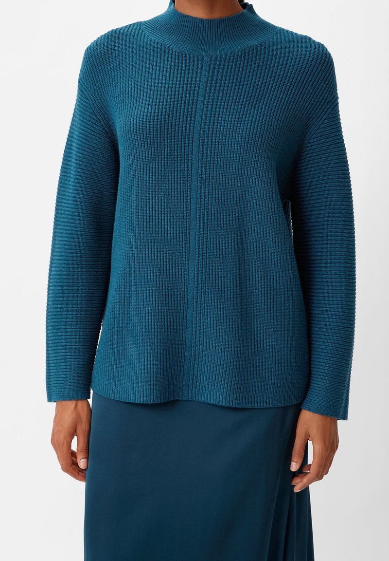 Pull en maille côtelée sarcelle avec col montant et manches longues porté avec une jupe assortie sarcelle, présenté des épaules aux hanches.