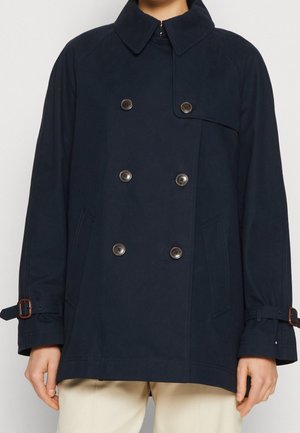 Cappotto doppiopetto blu navy con sei bottoni, ampio colletto, tasche laterali e cinturini con fibbia sulle maniche indossato sopra pantaloni beige.