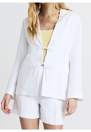 Kvinde iført en hvid blazer med to frontbind, matchende hvide shorts og en ribbet gul top underneath.