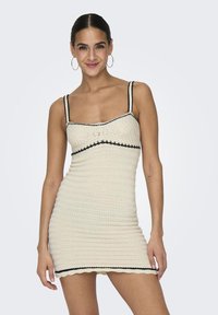 Knit dress made of cream-colored material with black trim. Features spaghetti straps and a fitted shape, with a scalloped hemline.