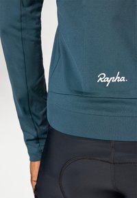 Ciklistična majica dolgem rokavu v teal barvi z oprijeto prileganjem in rebrastim robom, ki vsebuje bel 'Rapha' logo. V kombinaciji z črnimi podloženimi kolesarskimi kratkimi hlačami.
