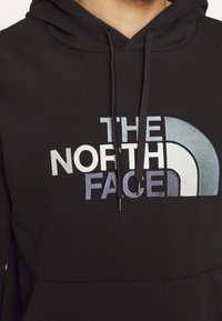 Černá mikina s kapucí a přední kapsou, na které je velké vyšívané logo v šedé, bílé a tyrkysové barvě. Měkká tkanina s šňůrkami na kapuci.