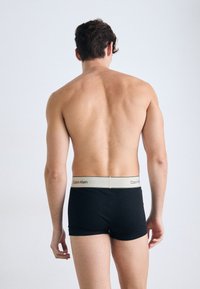 Schwarze Baumwollunterwäsche mit einem beigen Bund, der das Calvin Klein-Logo zeigt, von hinten betrachtet. Glatte Textur, taillierte Passform.