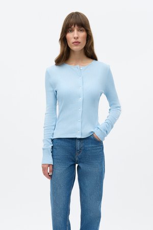 Világoskék bordázott kardigán hosszú ujjakkal, kerek nyakkivágással és apró gombokkal az elején, kék farmernadrággal párosítva.