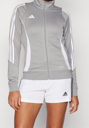 Kvinde iført en grå Adidas lynlåstrøje med hvide striber på ærmerne og hvide Adidas-shorts med sort logo.