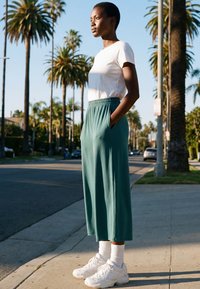 Young person standing on sidewalk with hands in pockets, wearing white t-shirt, green wide-leg pants, white socks, and sneakers, palm trees in background.