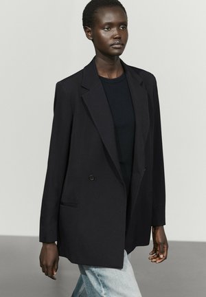 Femme à la peau foncée portant un blazer noir ample sur un haut noir et un jean bleu clair, debout devant un fond clair uni.