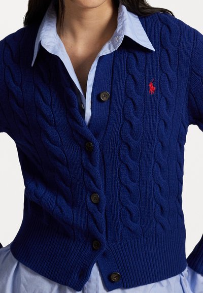 Blå kabelstrikket cardigan med knappfront, ribbkant og rødt brodert logo. Båret over en lyseblå skjorte med krage.