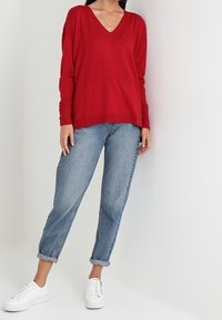Maglione rosso a collo a V, vestibilità rilassata, abbinato a jeans in denim a vita alta di lavaggio chiaro arrotolati. Sneakers bianche completano l'outfit. Look semplice e informale.