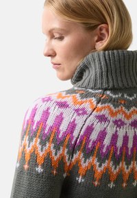 Žena s blonďatými vlasy, která má na sobě šedý rolák se světlými oranžovými, fialovými, bílými a šedými geometrickými vzory na ramenou.