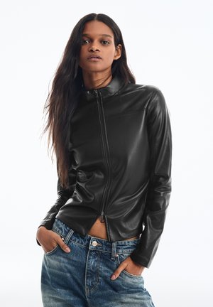 Femme aux longs cheveux foncés portant une veste en cuir noire zippée et un jean bleu, les mains dans les poches, sur un fond blanc uni.