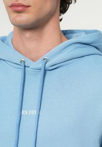 Közeli kép egy személyről, aki világoskék kapucnis pulóvert visel, rajta húzózsinórokkal és a mellrészen részben látható fehér szöveggel.