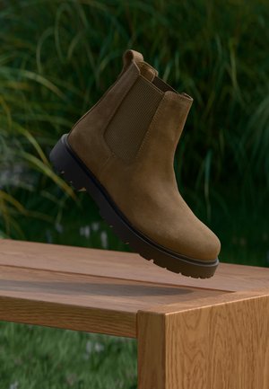 Brauner Chelsea-Stiefel aus Wildleder mit schwarzer Sohle, der auf der Kante eines Holztisches im Freien mit Grün im Hintergrund balanciert.