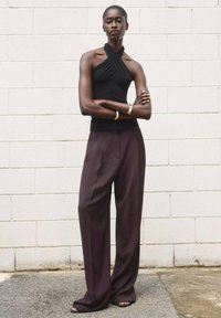 Black sleeveless top with a halter design, paired with loose, dark purple trousers, displayed against a neutral wall background.