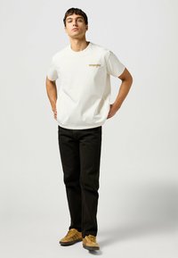 Camiseta de algodón blanca de manga corta y cuello redondo, con un logo dorado de "Wrangler" en el lado izquierdo del pecho, combinada con pantalones negros y zapatos beige.