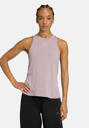 Femme aux cheveux tressés portant un haut de sport sans manches violet clair et un pantalon noir, debout avec une main sur la hanche devant un fond uni.