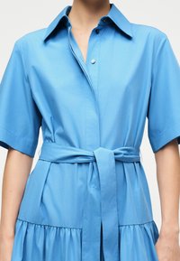 Φόρεμα πουκάμισο σε ανοιχτό γαλάζιο χρώμα με κοντά μανίκια, γιακά, κουμπωτό μπροστινό μέρος και ζώνη που δένει στη μέση.