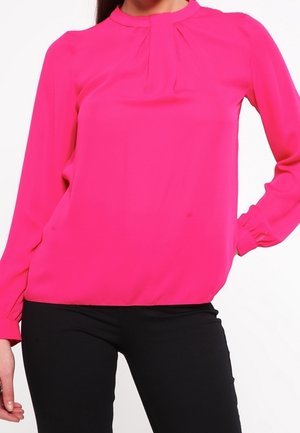 Femme portant un chemisier rose vif à manches longues avec des plis au niveau de l'encolure et un pantalon noir ajusté, debout avec une main sur la hanche.