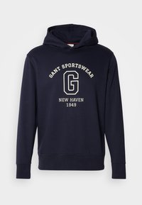 Sweat à capuche bleu marine avec « GANT SPORTSWEAR », un grand « G » et « NEW HAVEN 1949 » imprimés en blanc sur le devant.