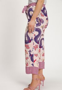 De stoffen broek heeft een roze basis met draken- en bloemmotieven in blauw, rood en goud. Een ceintuur in de taille en een patroon aan de zoom maken het ontwerp compleet.