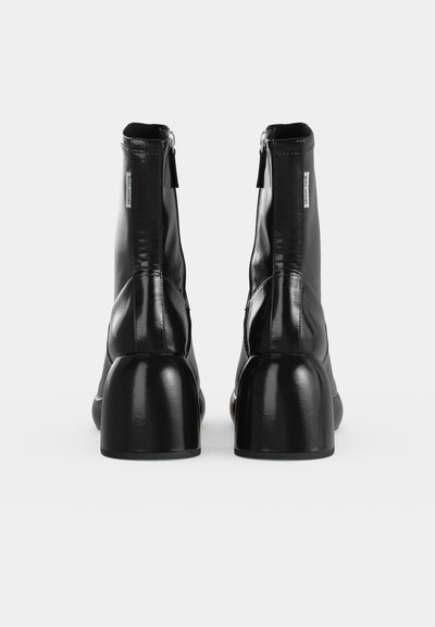 Paire de bottines noires en cuir brillant avec talons carrés et fermetures éclair à l'arrière, vues de dos sur fond blanc.