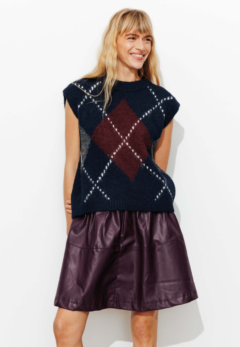 Pull sans manches bleu marine avec motif argyle en bordeaux et blanc, associé à une jupe en cuir violette foncée taille haute. Texture douce.